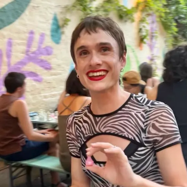Photograph of chess coach Niki. She's in front of a chess board with a pink pawn in her hand. There are others playing chess in the background and partial view of a mural on a brick wall.