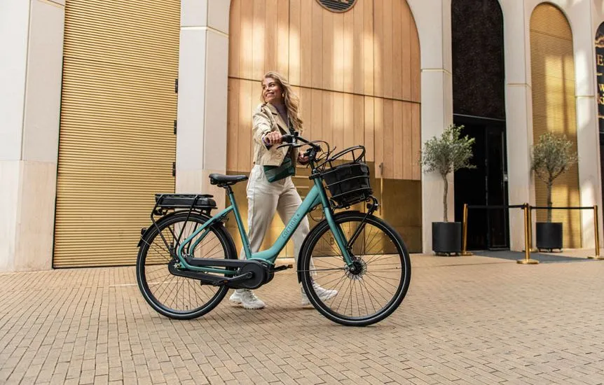 Vrouw lopend op straat met een Cortina E-Common Jade Green elektrische damesfiets aan de hand met voorop een fietsmandje 